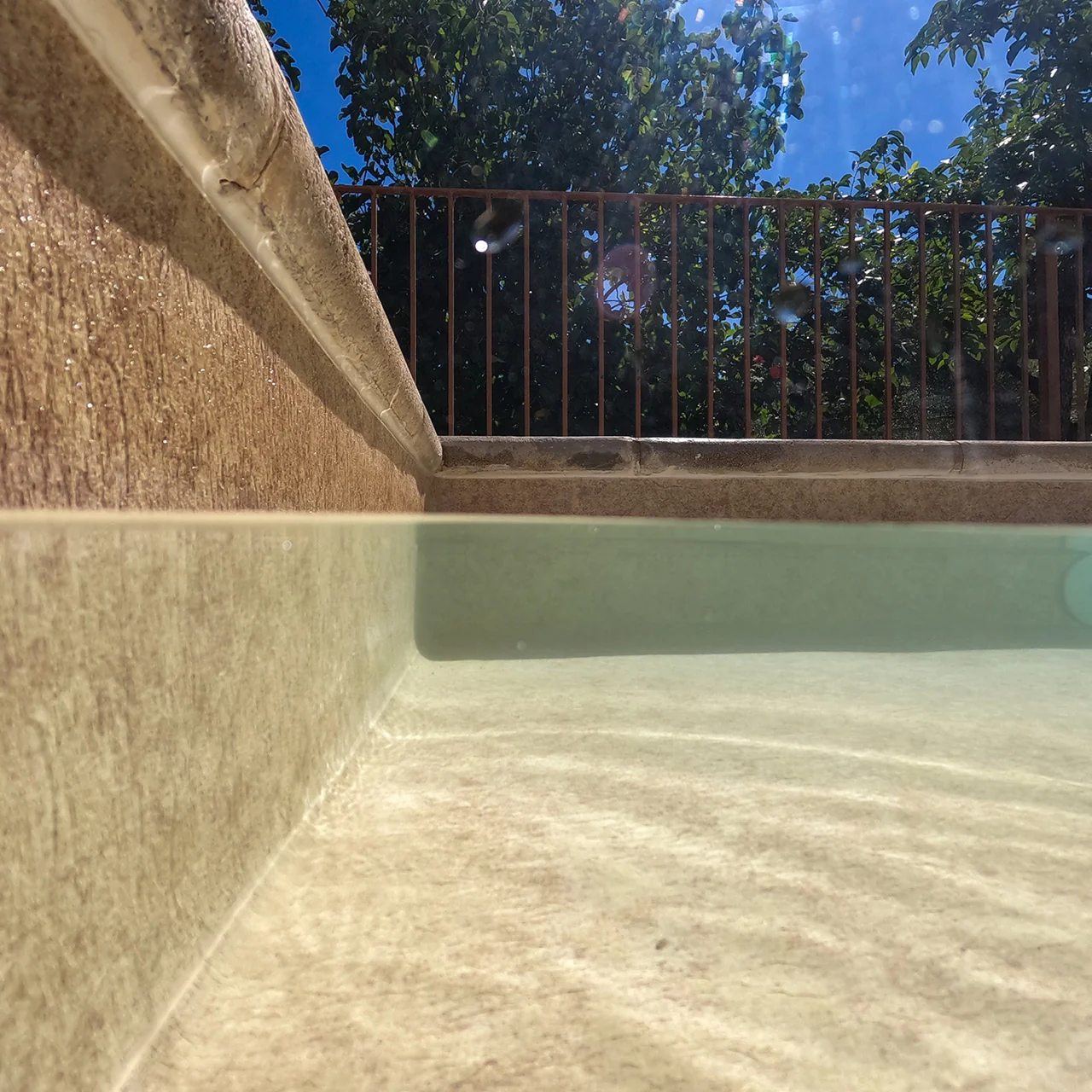 Vue au ras de l’eau d’une piscine avec membrane armée BWT Be BRANE 3D Surface Beach Sand, teinte sable crème et margelle en pierre claire