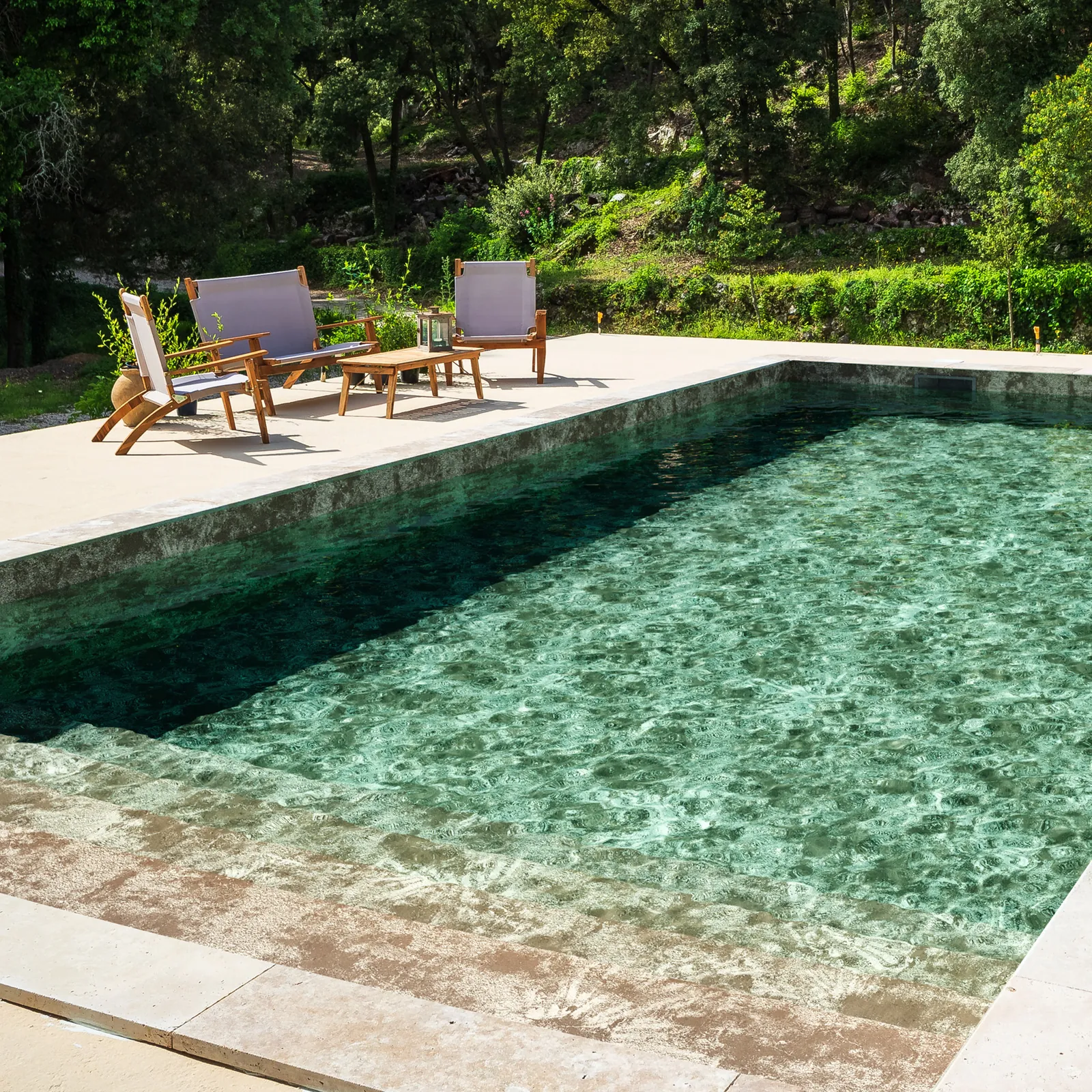Piscine avec membrane armée BWT Be BRANE 3D Surface Coral Reef Sand, eau vert émeraude et margelle en pierre sable face à une forêt méditerranéenne